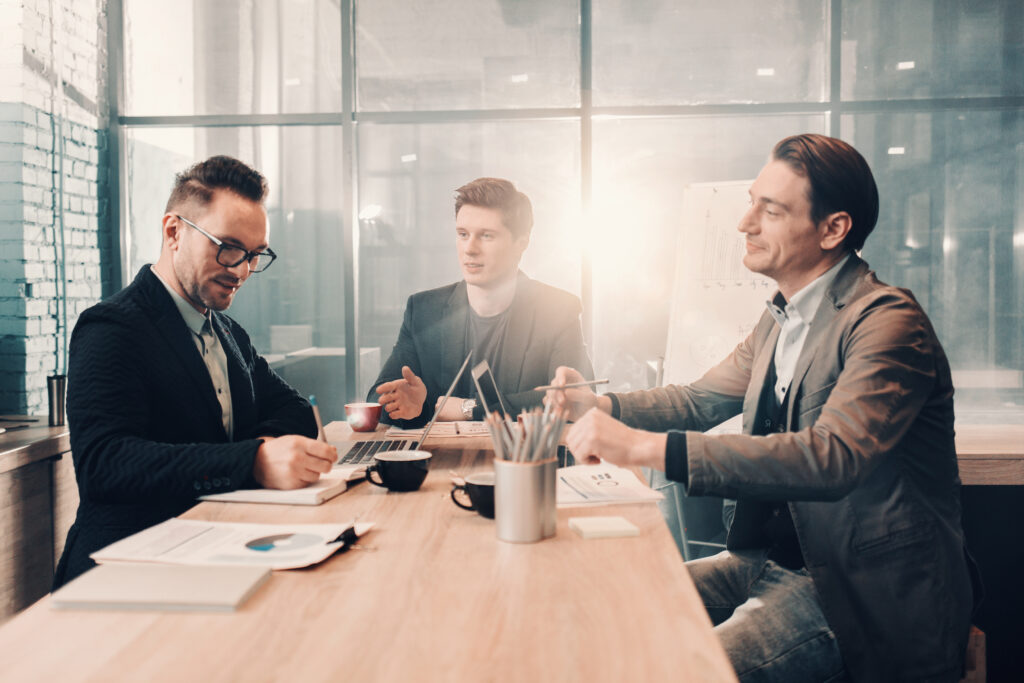 Portrait of smiling bearded man writing in copybook while speaking with happy partners. They situating at desk. Communication and business concept
